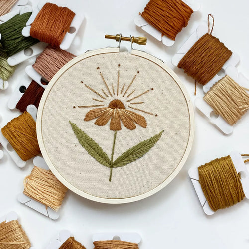 Embroidery hoop holds tan fabric stitched with warm sunflower or daisy-styled flower, surrounded by warm-toned threads.