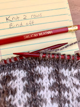 Load image into Gallery viewer, A red pencil with the words &quot;Sweatah Weathah&quot; in gold lettering lies atop a notepad with knit pattern instructions, next to a gray and white knit project on the needles.
