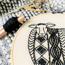 Load image into Gallery viewer, The corner of an embroidery hoop and print in black embroidery thread, of a sheep wearing a cable knit sweater. Scissors and embroidery thread lie nearby.

