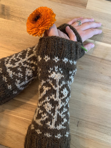 A pair of hands hold an orange calendula blossom and wear a pair of brown knit armwarmers almost to the elbow, with ribbing at either end and a white colorwork motif of vines and flowers.
