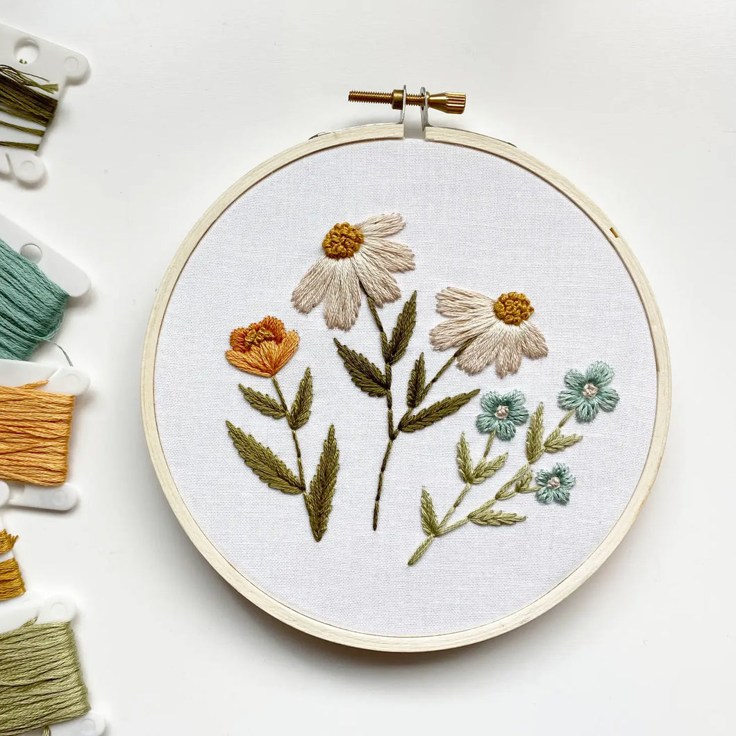 An embroidery hoop holds white fabric stitched with four different flower designs in soft shades of white, gold and blue.