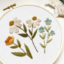 Load image into Gallery viewer, An embroidery hoop holds white fabric stitched with four different flower designs in soft shades of white, gold and blue.
