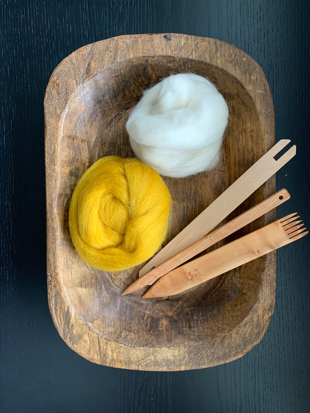 A carved wood bowl holds wood tapestry weaving tools and two bumps of roving in white and mustard yellow.